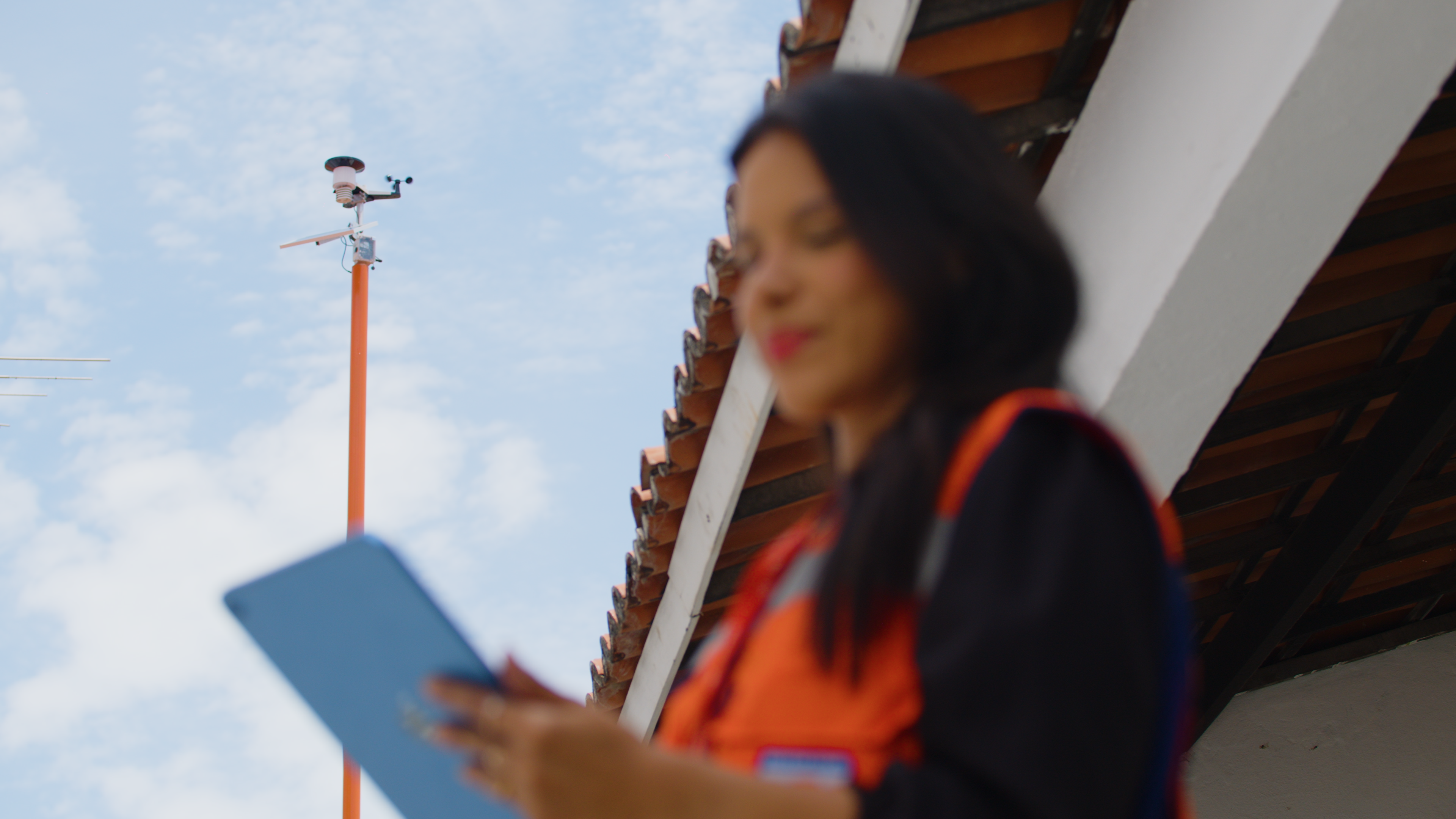 Mulher segura um tablet. Ao fundo aparece uma estação metereológica